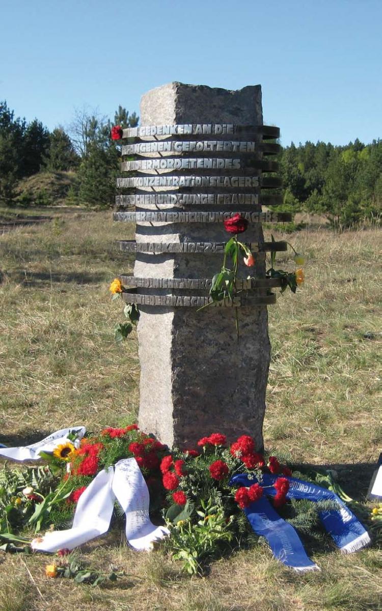 Säulenförmiger Gedenkstein mit geschmiedeter Schrift, davor Blumengestecke